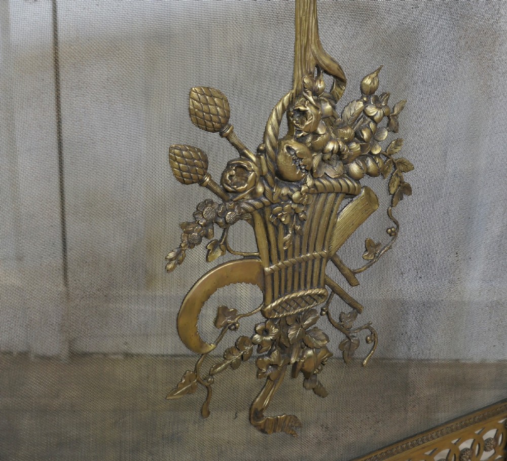 A freestanding brass fire guard featuring a central ribbon bow and floral basket relief mounted on fine wire mesh with scroll feet. View VII