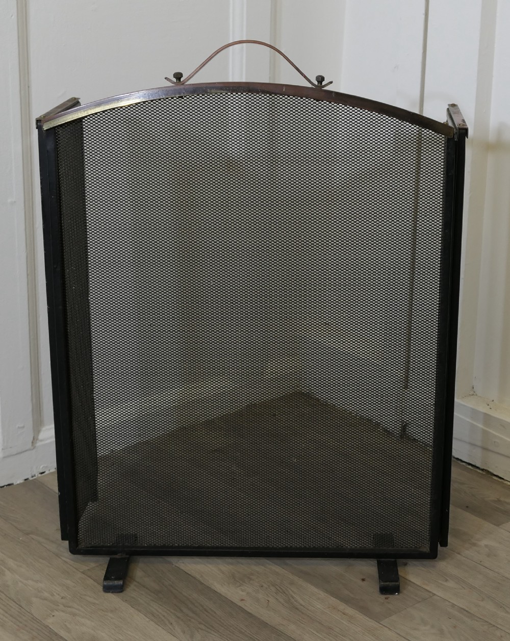A folding black metal wire mesh fire guard featuring an arched copper top rail with a central carrying handle.