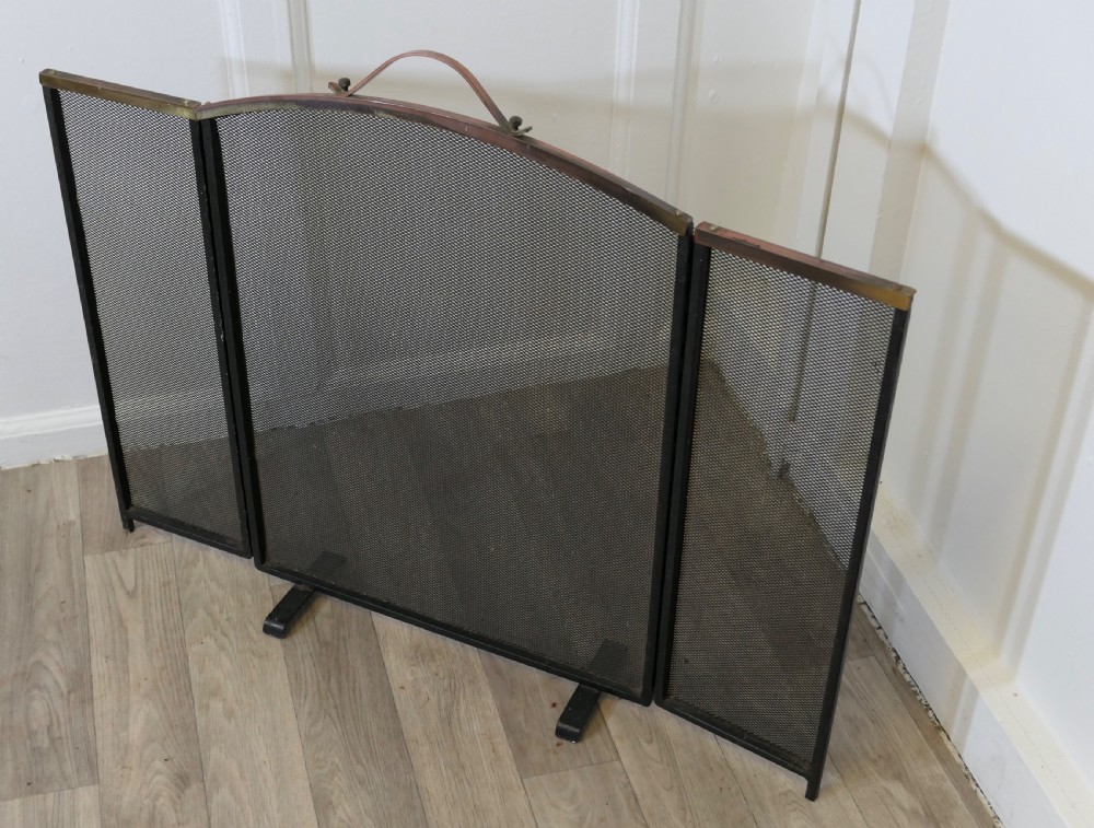A folding black metal wire mesh fire guard featuring an arched copper top rail with a central carrying handle. View V