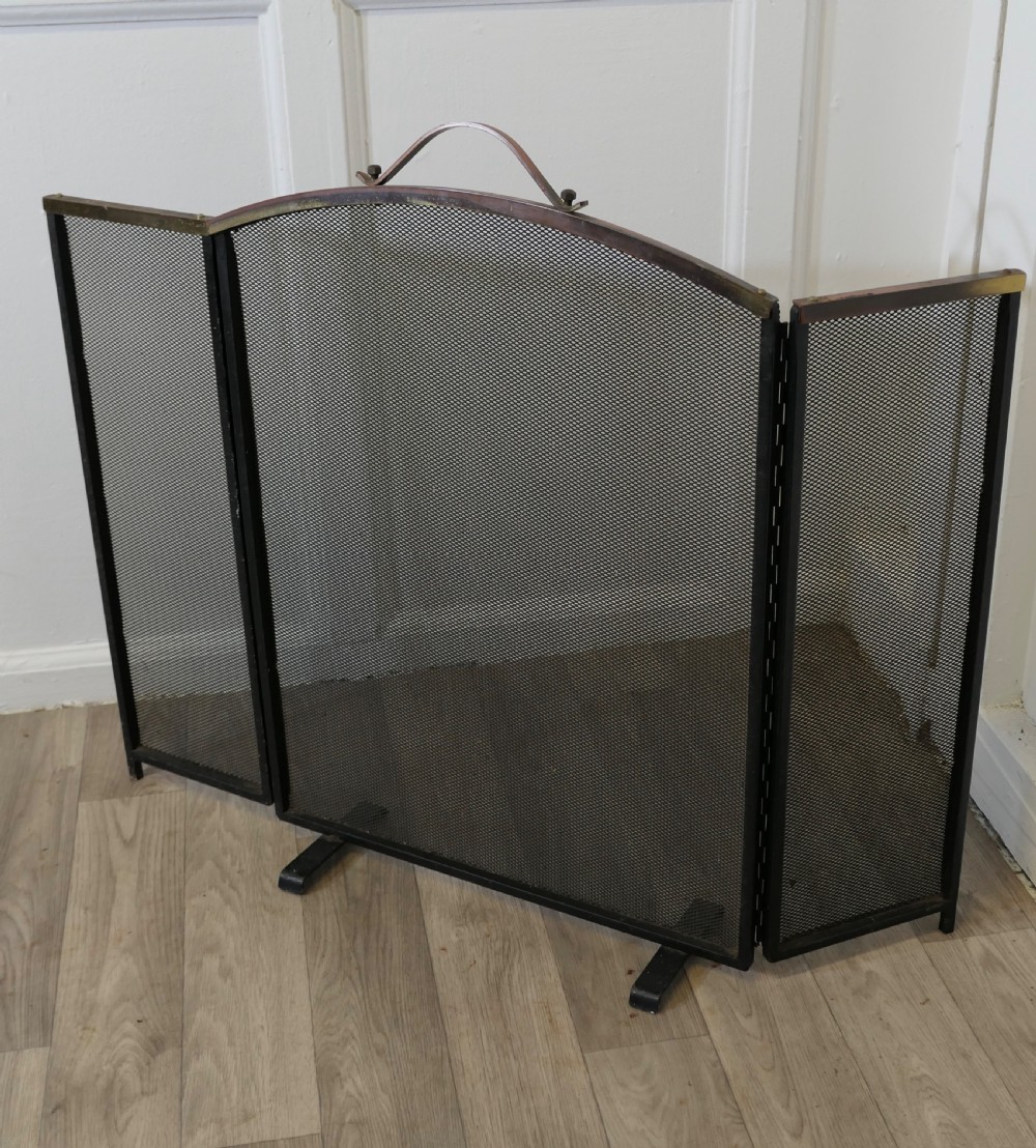 A folding black metal wire mesh fire guard featuring an arched copper top rail with a central carrying handle. View VI