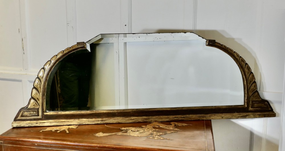 A wide Art Deco overmantel mirror featuring a low horizontal frame with dark wood moulding and gold gilded shoulders carved in a stylised leaf pattern.