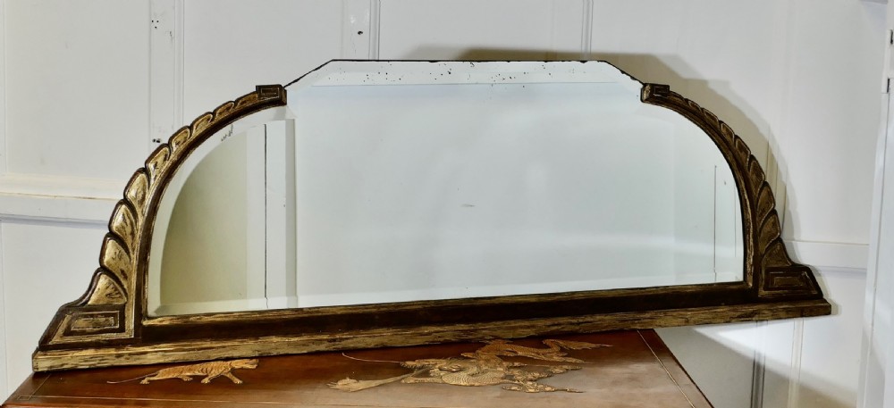 A wide Art Deco overmantel mirror featuring a low horizontal frame with dark wood moulding and gold gilded shoulders carved in a stylised leaf pattern. View II