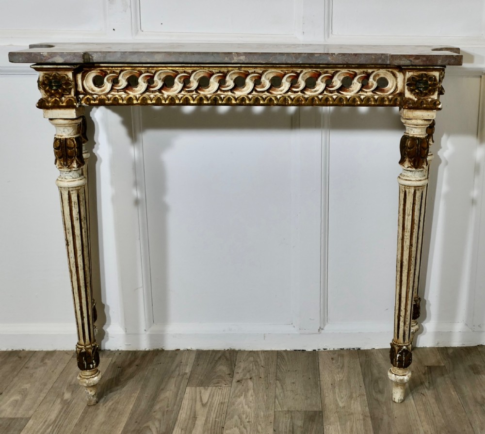 A French Neoclassical style console table featuring a grey marble top, carved pierced frieze, and cream painted tapered fluted legs with gilded accents.