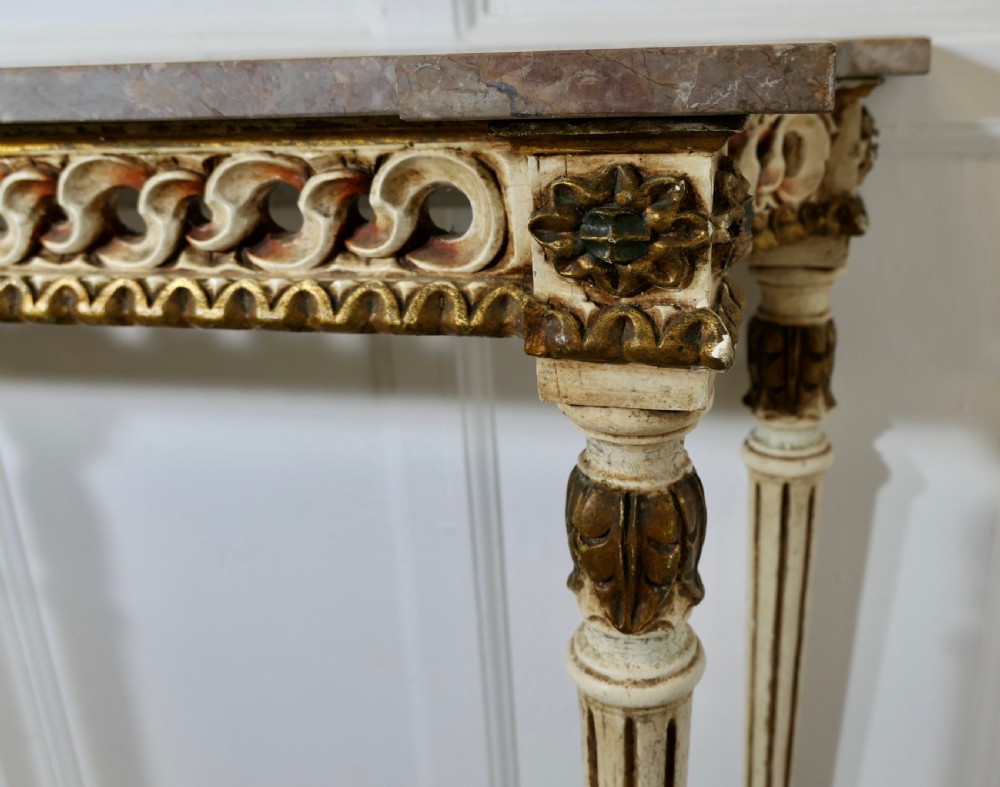A French Neoclassical style console table featuring a grey marble top, carved pierced frieze, and cream painted tapered fluted legs with gilded accents. View III