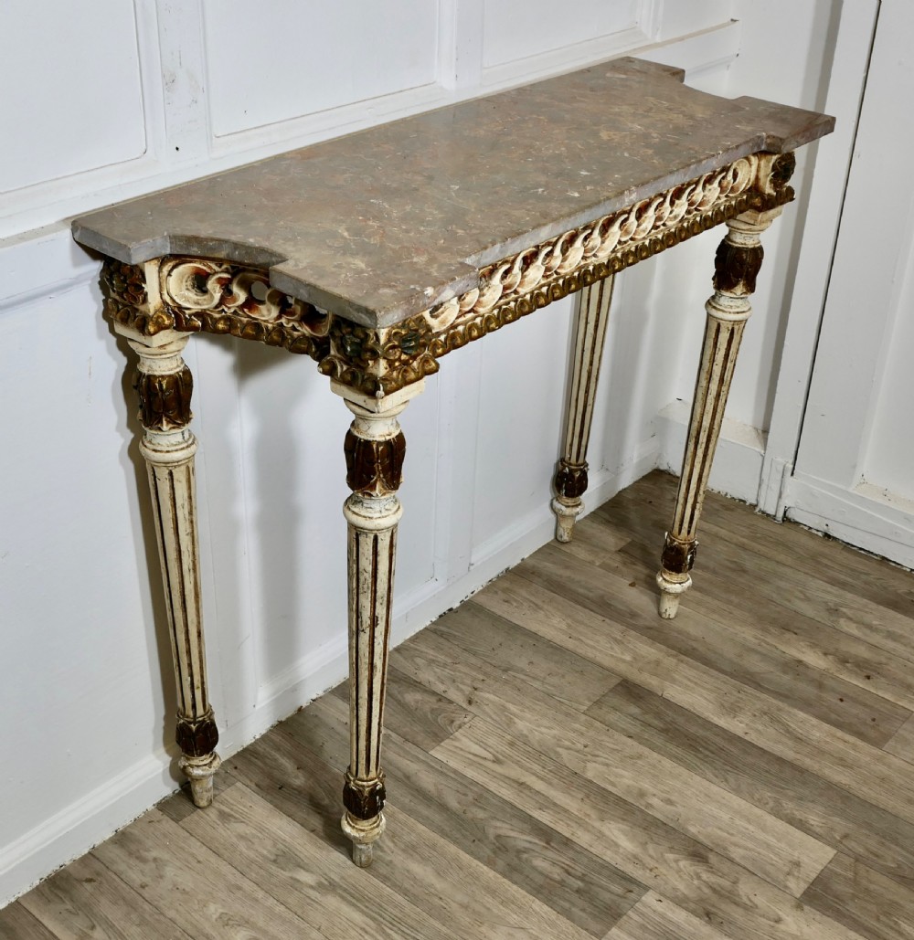 A French Neoclassical style console table featuring a grey marble top, carved pierced frieze, and cream painted tapered fluted legs with gilded accents. View V