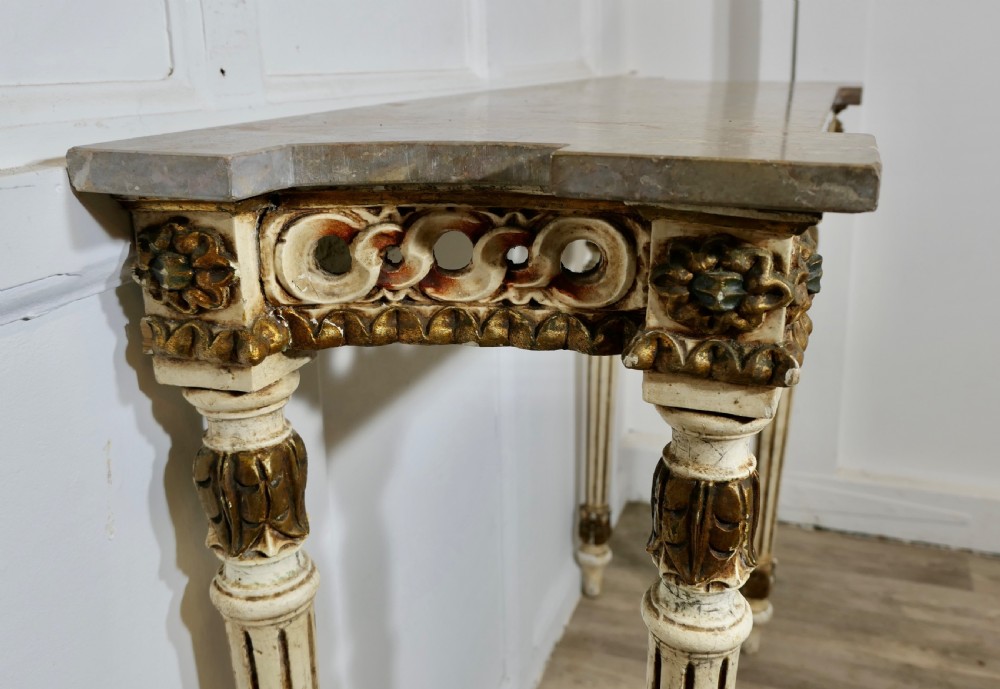 A French Neoclassical style console table featuring a grey marble top, carved pierced frieze, and cream painted tapered fluted legs with gilded accents. View VI
