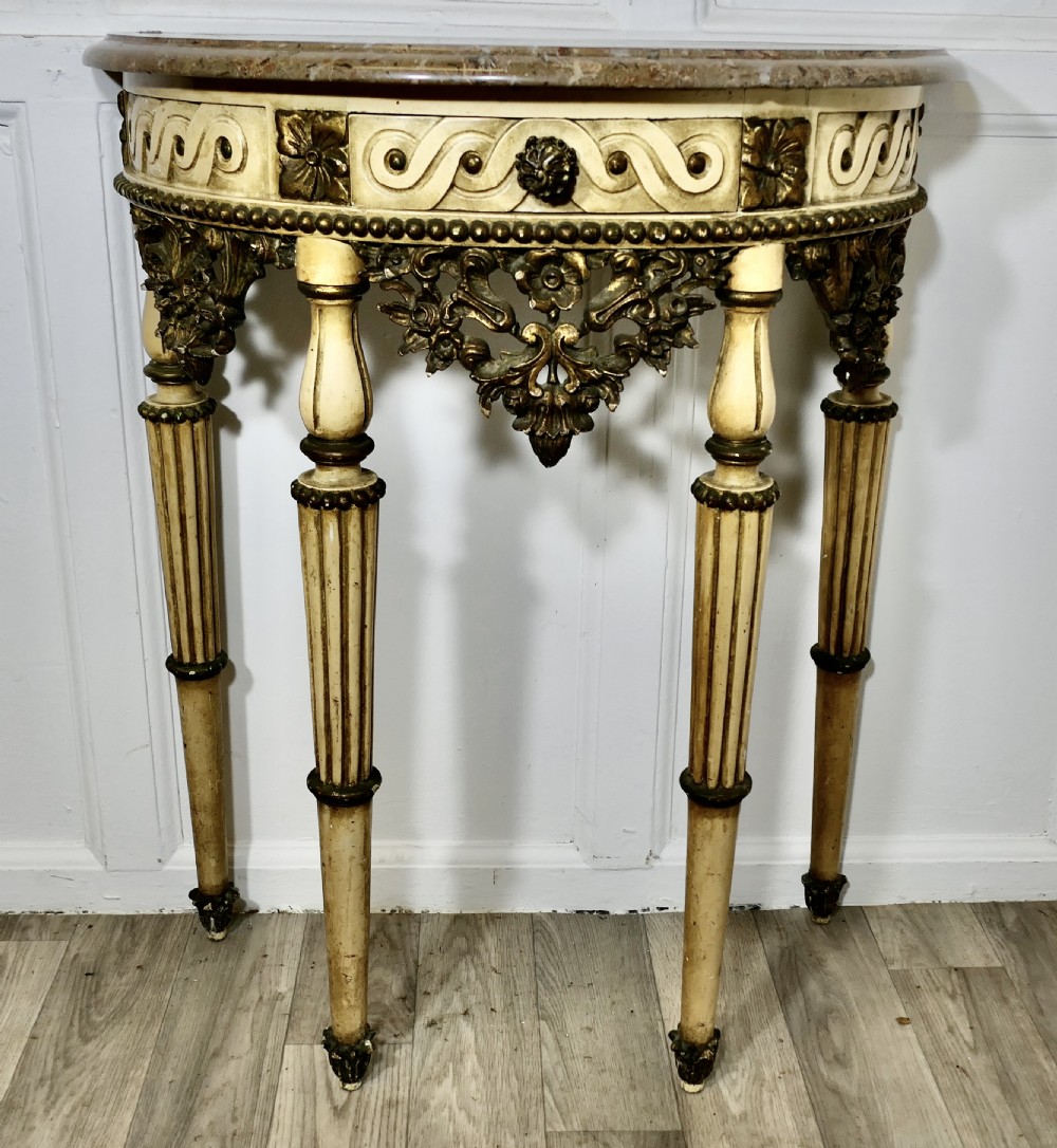 A semi-circular demilune console table featuring a marble top, a painted cream frame with gold leaf highlights, and carved fluted legs.
