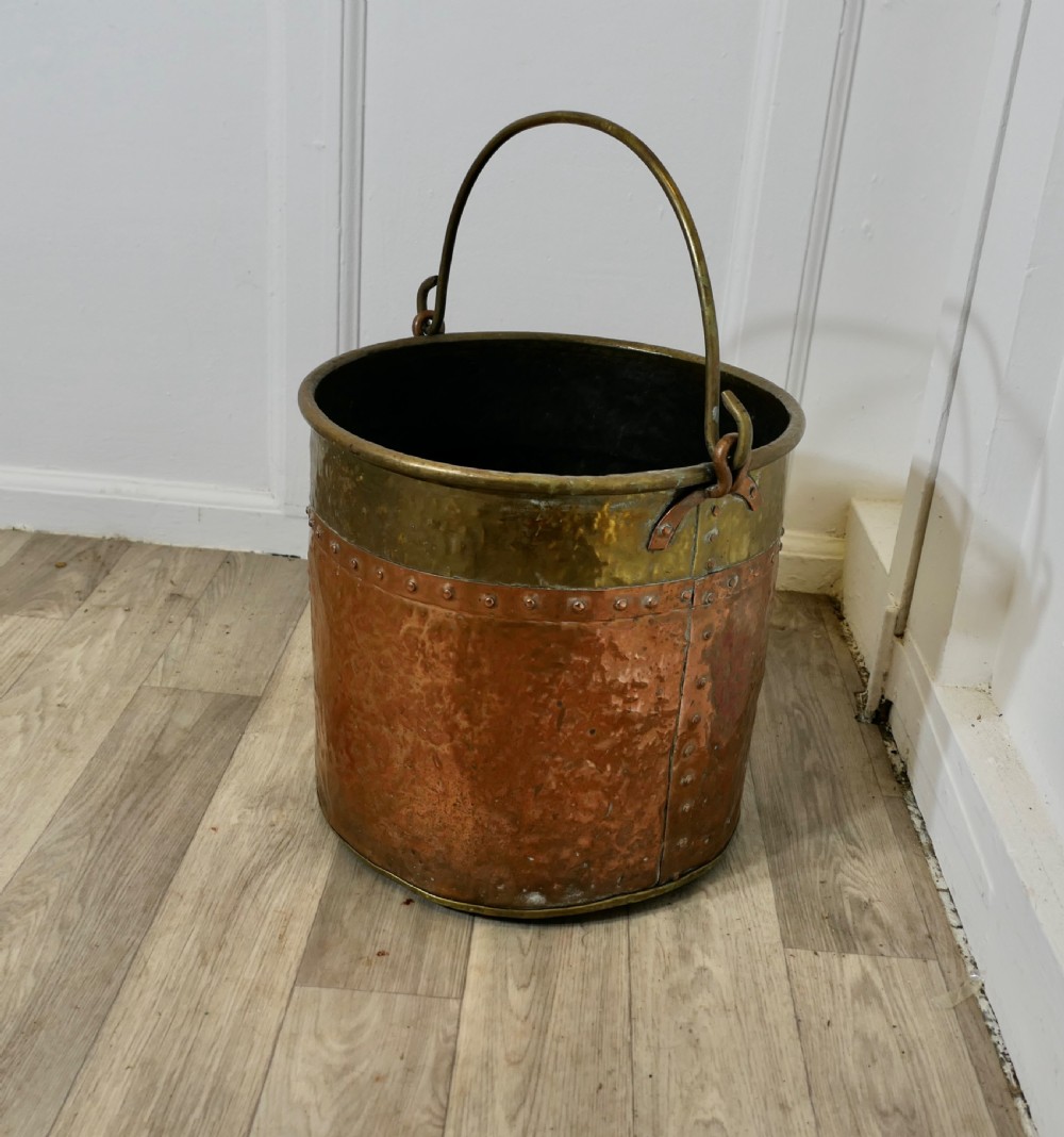 A cylindrical hand-hammered copper bucket featuring a wide brass top collar, riveted seam, and a curved brass swing handle. View X