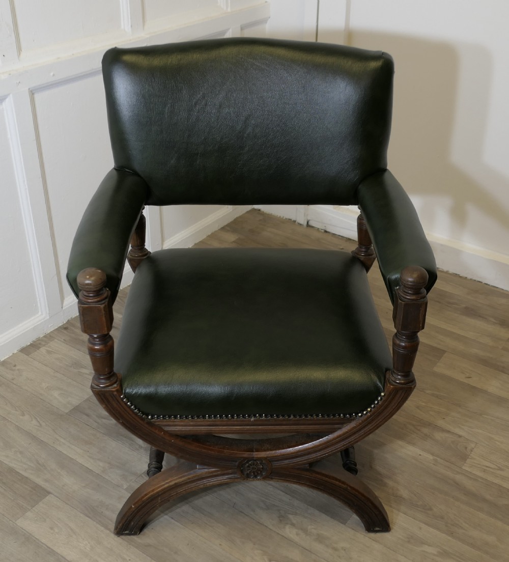 A 19th-century desk chair featuring dark green leather upholstery, brass stud detailing, turned arm supports, and a carved wooden X-frame base.