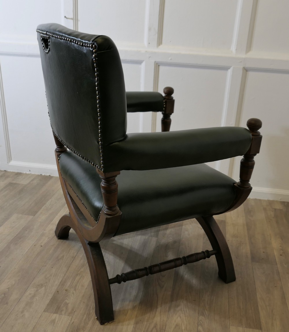 A 19th-century desk chair featuring dark green leather upholstery, brass stud detailing, turned arm supports, and a carved wooden X-frame base. View VI