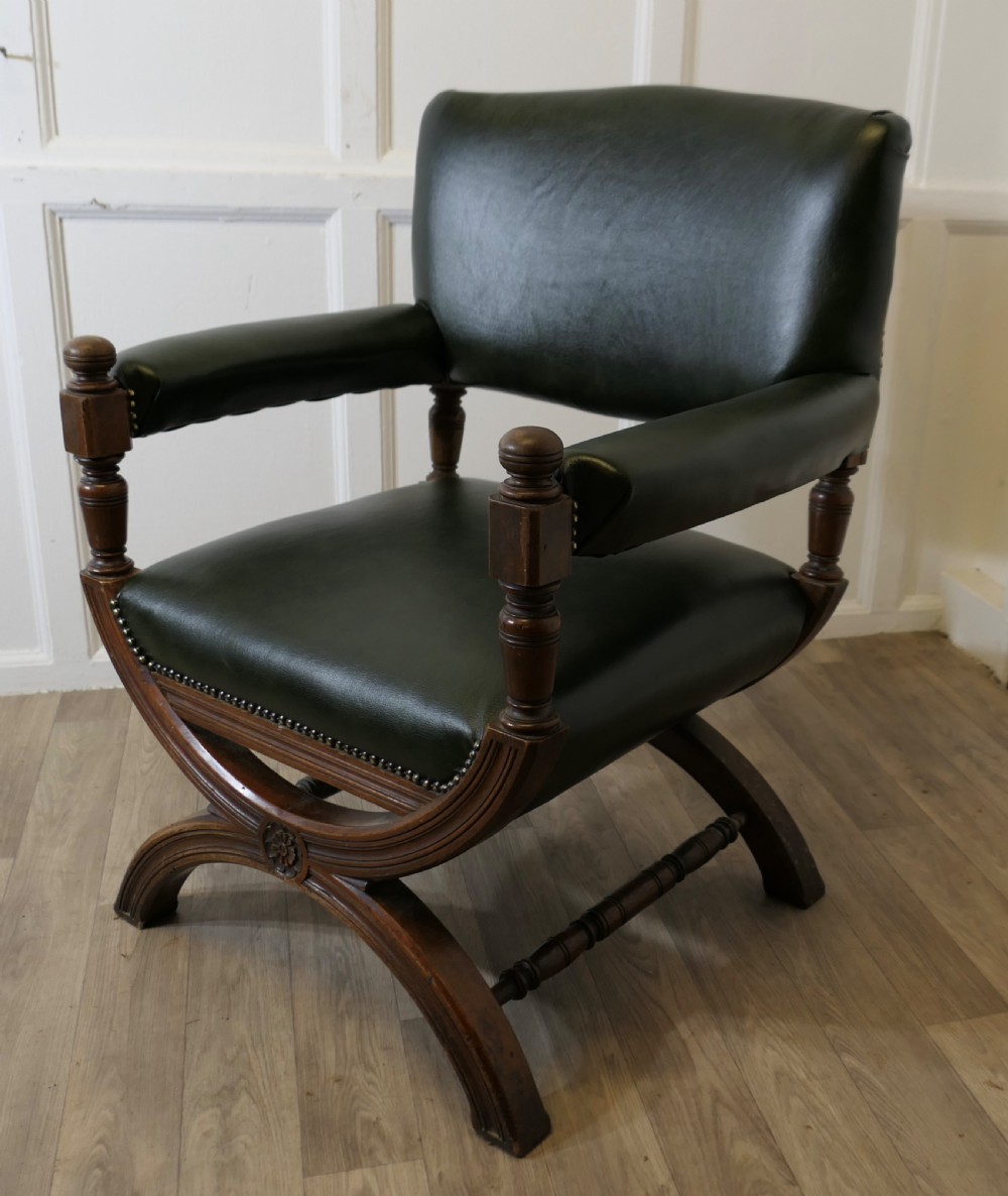 A 19th-century desk chair featuring dark green leather upholstery, brass stud detailing, turned arm supports, and a carved wooden X-frame base. View X