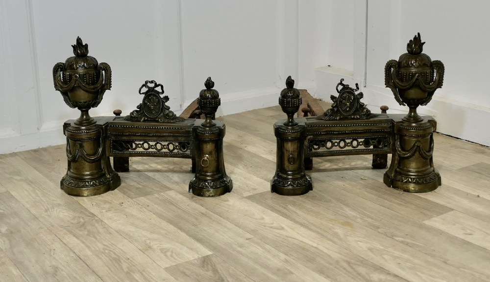 A pair of 18th-century French Neoclassical brass chenets featuring flame-topped urns, drapery swags, and pierced friezes supported on circular bases.
