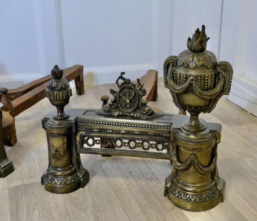 A pair of 18th-century French Neoclassical brass chenets featuring flame-topped urns, drapery swags, and pierced friezes supported on circular bases. View V