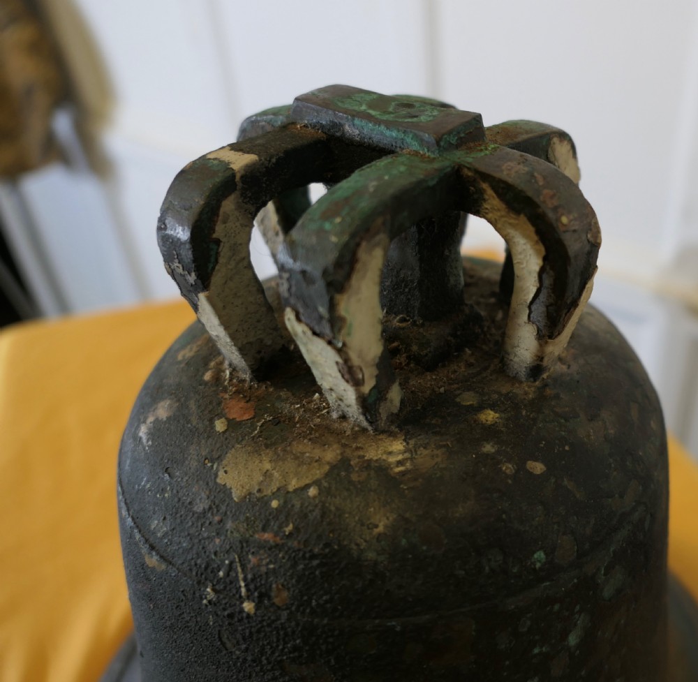 A heavy cast bronze tower bell with a dark weathered patina, white paint remnants, and arched crown loops at the top. View IV