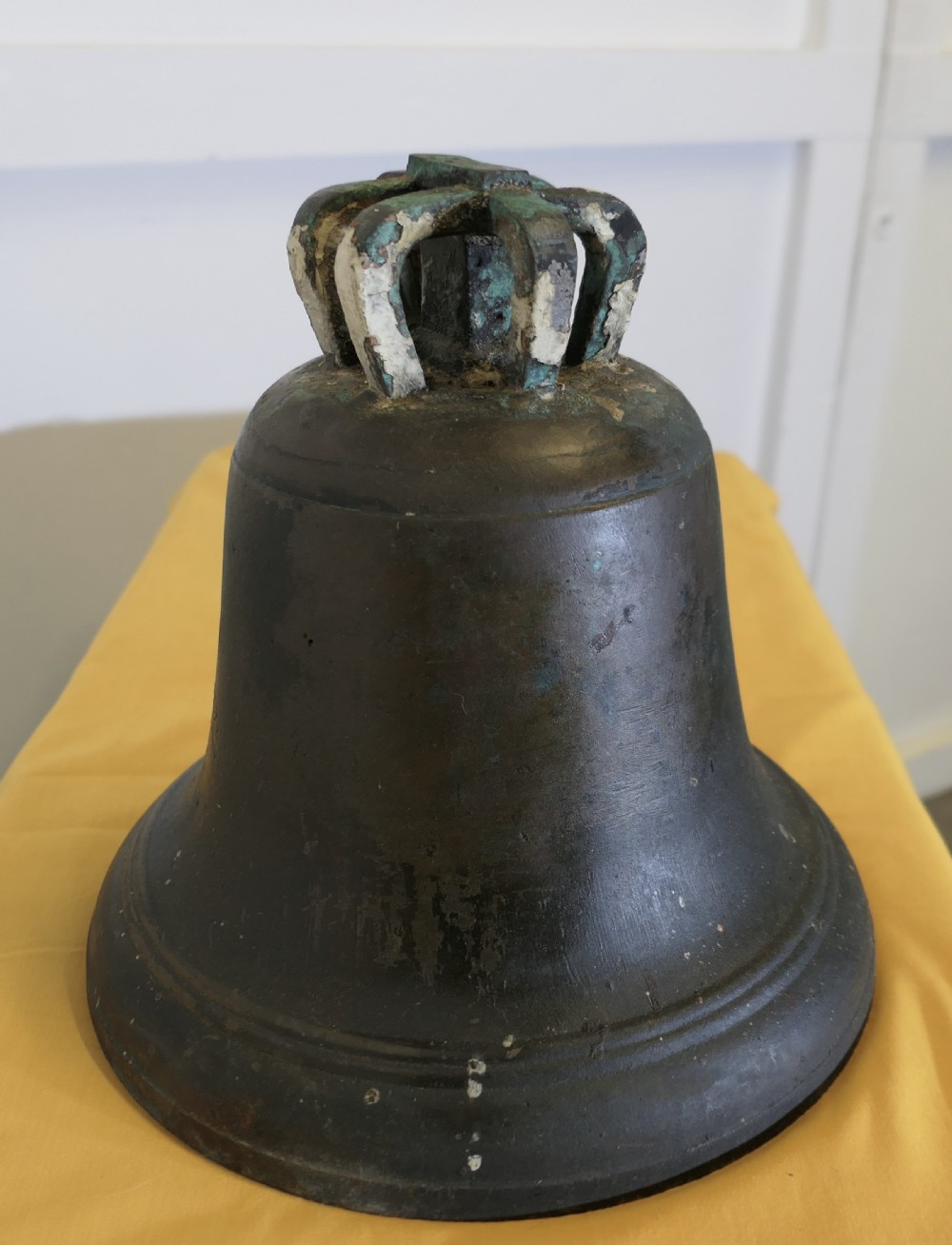 A heavy cast bronze tower bell with a dark weathered patina, white paint remnants, and arched crown loops at the top. View V