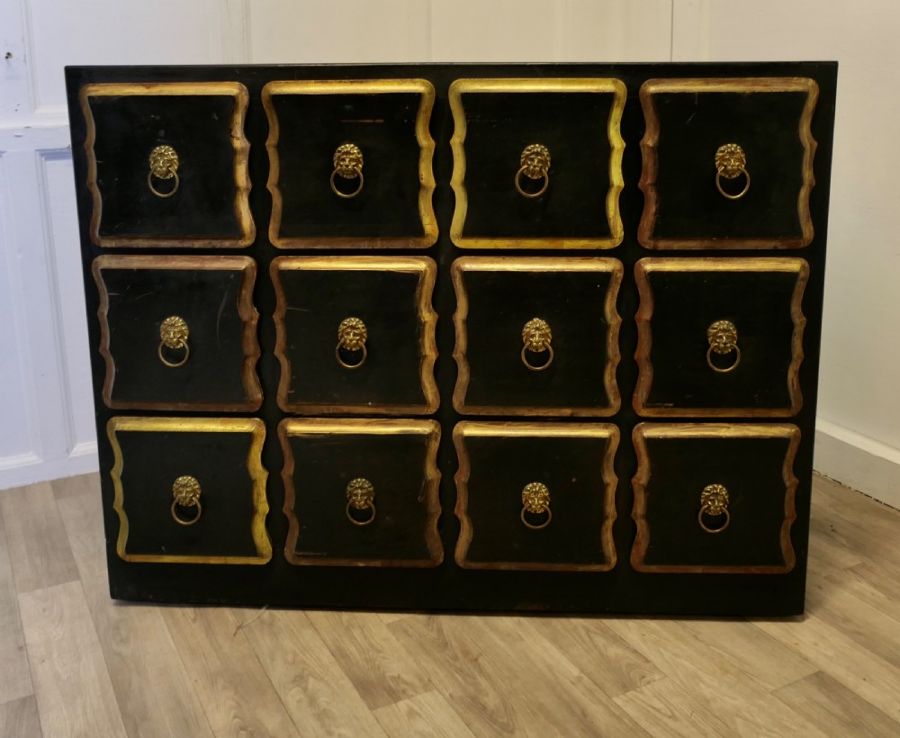A black ebonised wooden chest featuring twelve small drawers detailed with contoured gold leaf borders and brass lion mask ring pulls.