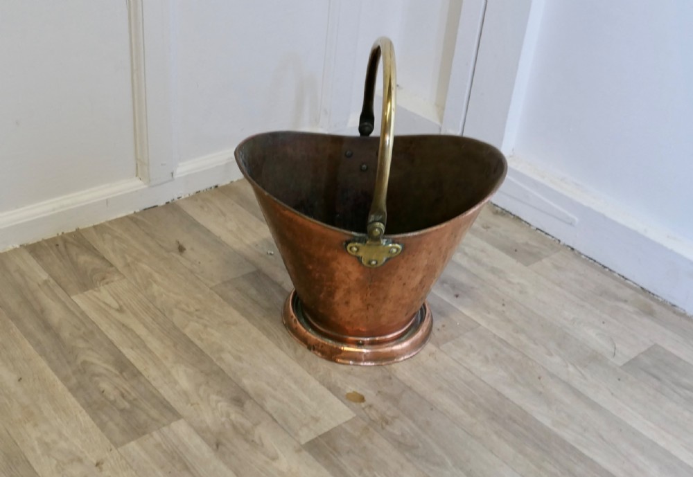 An antique hand-hammered copper helmet coal scuttle featuring a fixed brass overhead handle and riveted mounts. View II