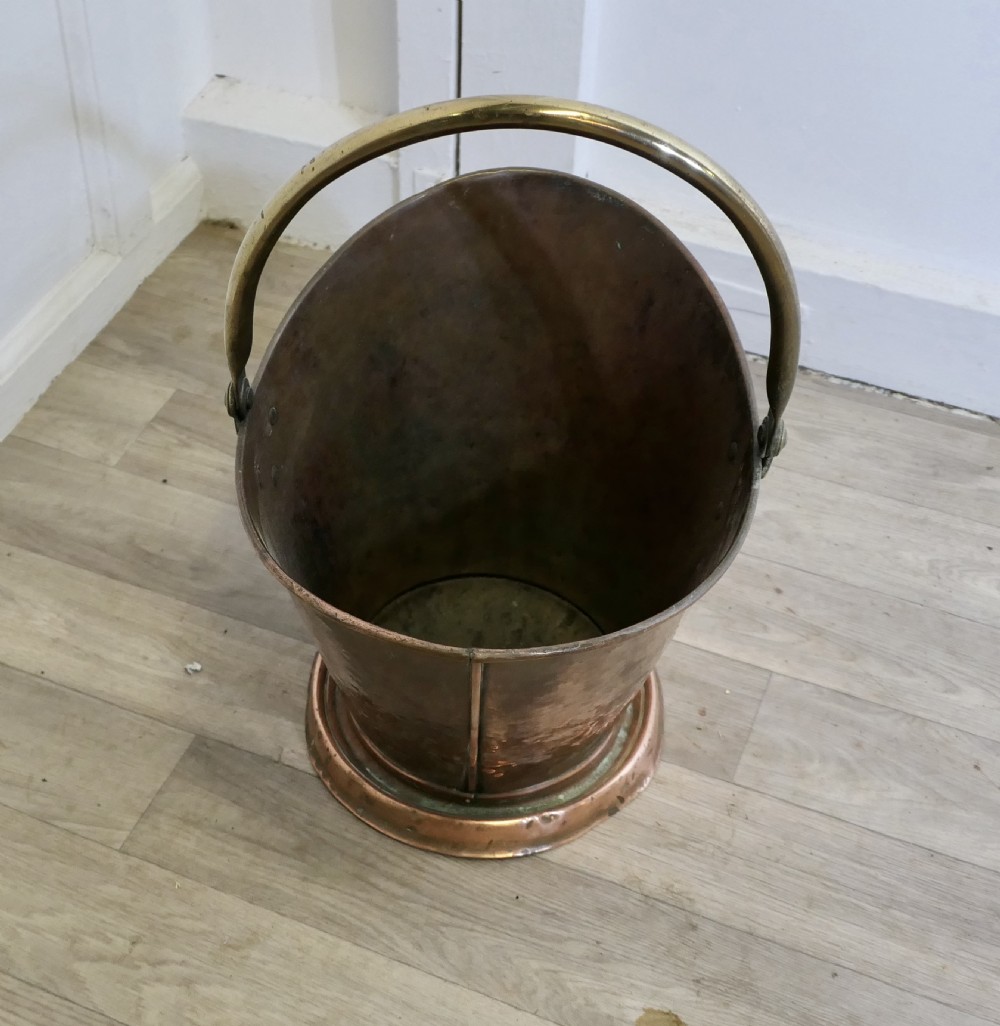 An antique hand-hammered copper helmet coal scuttle featuring a fixed brass overhead handle and riveted mounts. View IV