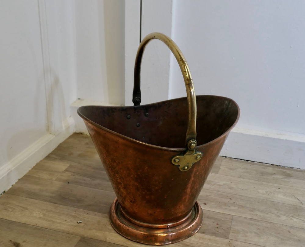 An antique hand-hammered copper helmet coal scuttle featuring a fixed brass overhead handle and riveted mounts. View V