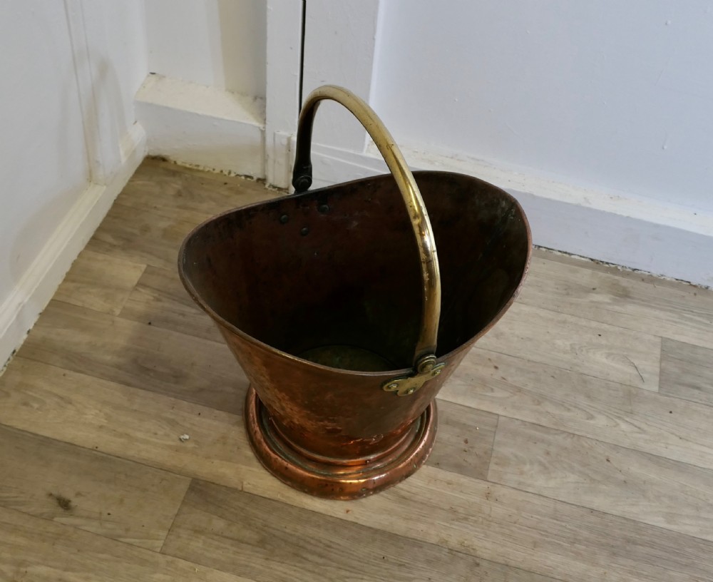 An antique hand-hammered copper helmet coal scuttle featuring a fixed brass overhead handle and riveted mounts. View VI