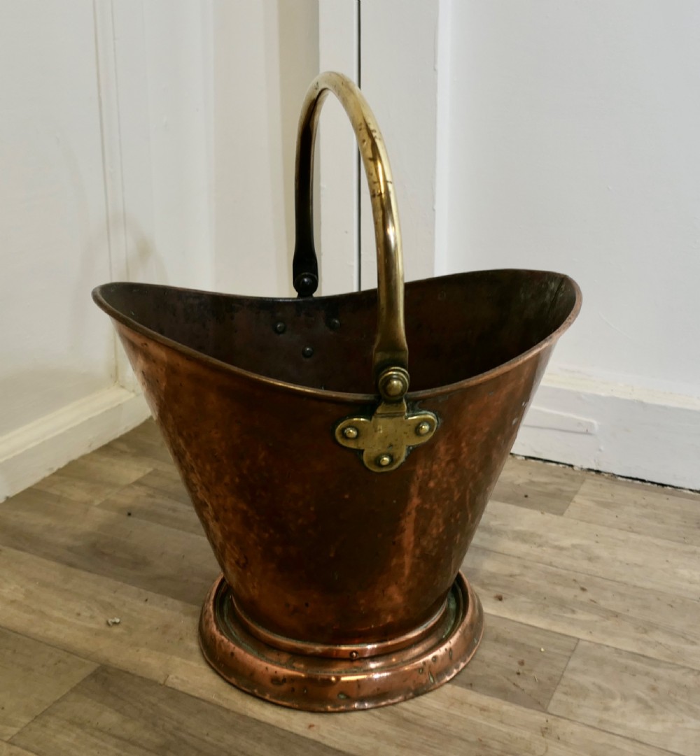 An antique hand-hammered copper helmet coal scuttle featuring a fixed brass overhead handle and riveted mounts. View VII