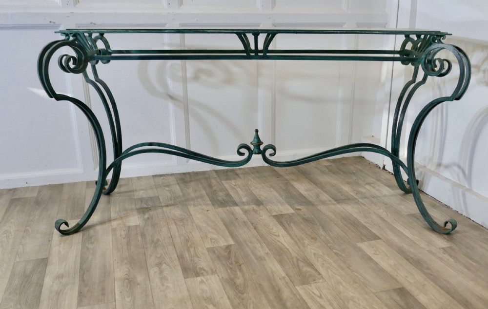 A green-painted wrought iron console table featuring curved scroll legs, a brass central finial, and a clear tabletop surface.