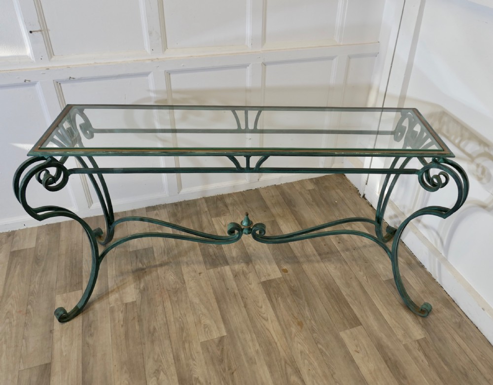 A green-painted wrought iron console table featuring curved scroll legs, a brass central finial, and a clear tabletop surface. View V