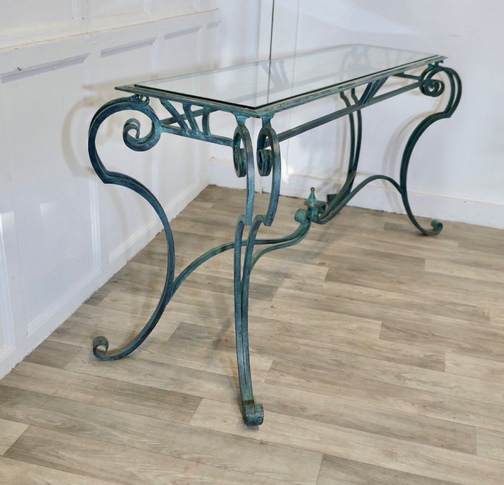 A green-painted wrought iron console table featuring curved scroll legs, a brass central finial, and a clear tabletop surface. View VII