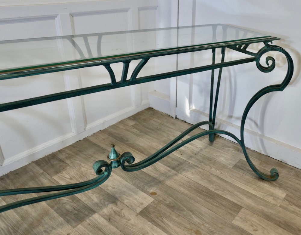 A green-painted wrought iron console table featuring curved scroll legs, a brass central finial, and a clear tabletop surface. View IX