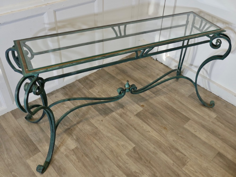 A green-painted wrought iron console table featuring curved scroll legs, a brass central finial, and a clear tabletop surface. View X