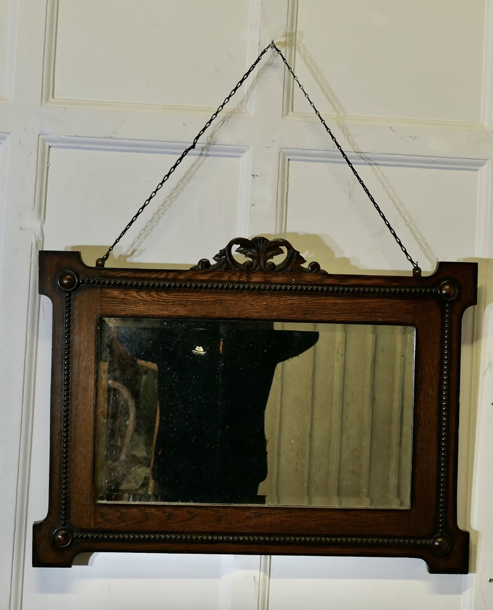 A horizontal rectangular dark oak framed mirror featuring a top carved crest, beaded inner moulding, and an attached hanging chain.