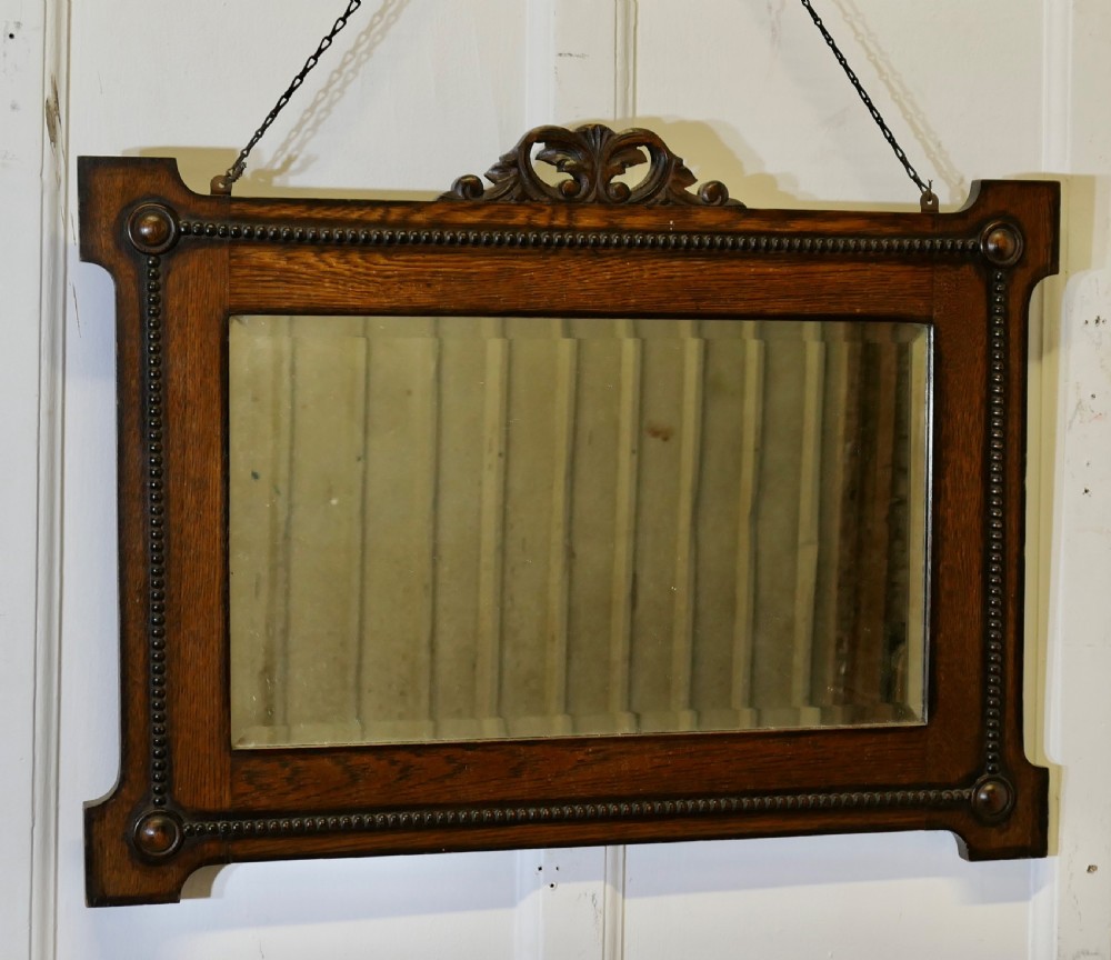 A horizontal rectangular dark oak framed mirror featuring a top carved crest, beaded inner moulding, and an attached hanging chain. View II