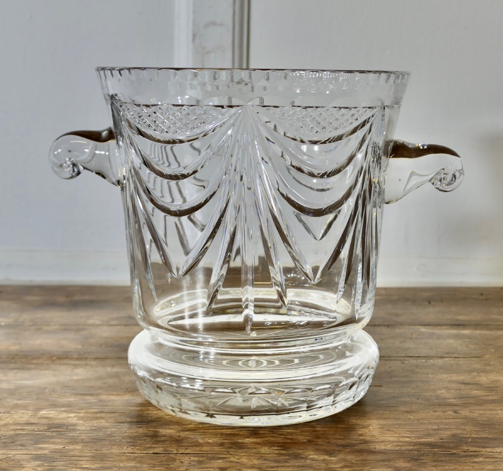 A heavy clear cut crystal champagne ice bucket featuring geometric drapery cuts, two applied scroll handles, and a stepped circular base.