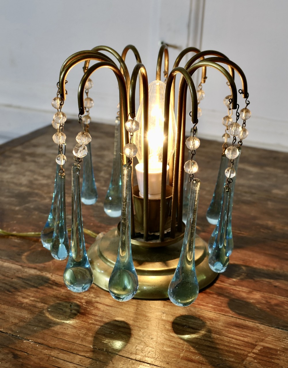 A small brass table lamp with curved radiating arms hanging clear crystal beads and light blue teardrop glass prisms around a central illuminated bulb. View IV