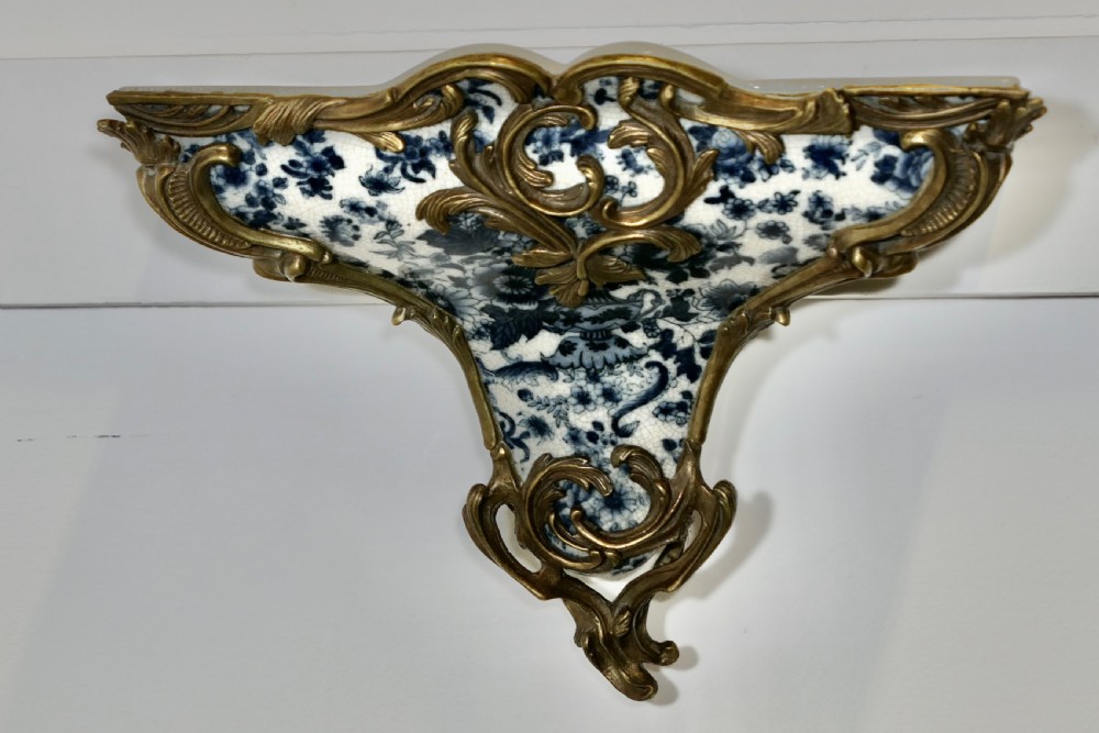 A Rococo revival wall bracket featuring blue and white floral glazed ceramic enclosed in ornamental gilt bronze foliage scrollwork.