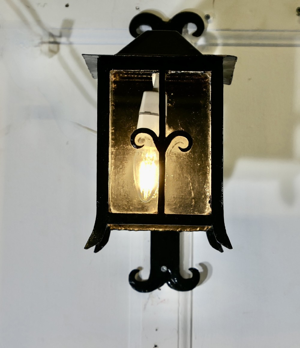 A black wrought iron wall lantern featuring clear glass panels, decorative scrollwork accents, and an illuminated flame-shaped bulb.