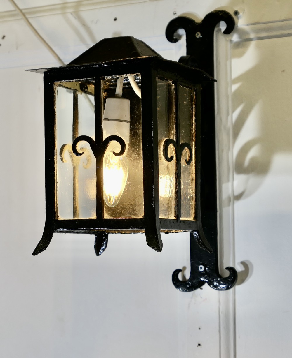 A black wrought iron wall lantern featuring clear glass panels, decorative scrollwork accents, and an illuminated flame-shaped bulb. View II