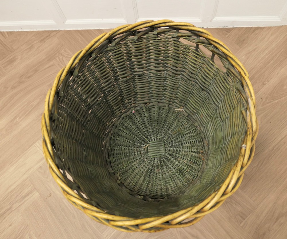 A large circular woven willow log basket with a braided rim resting on a wooden floor. View XI