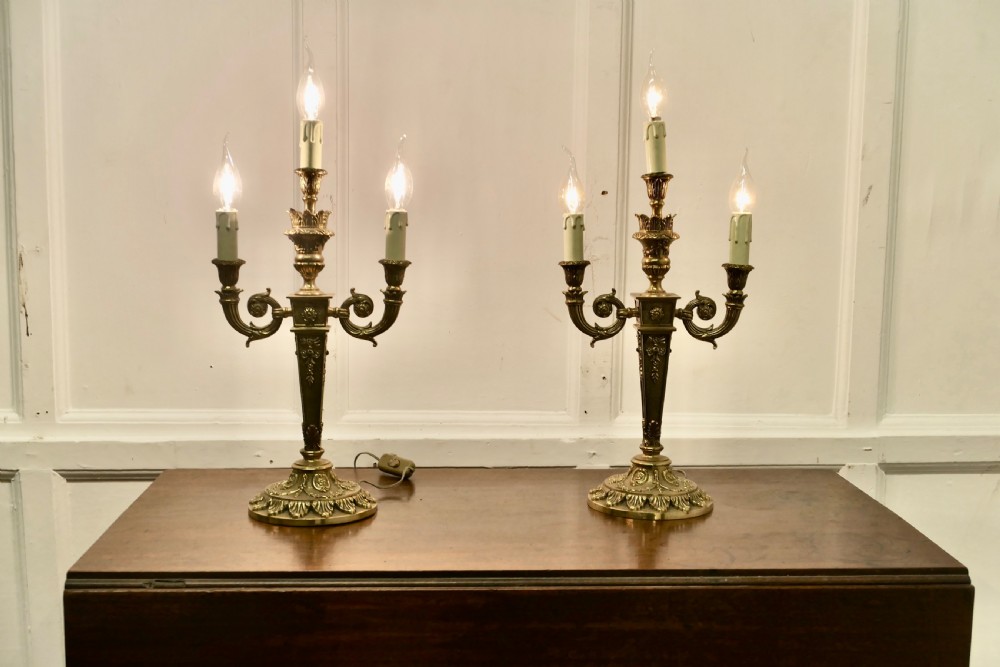 A matching pair of three-branch ornate brass electric candelabra table lamps featuring flame-shaped light bulbs and scrolled arms. View II