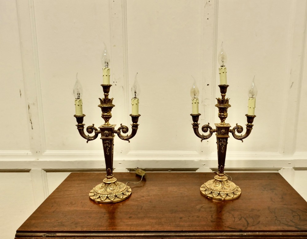 A matching pair of three-branch ornate brass electric candelabra table lamps featuring flame-shaped light bulbs and scrolled arms. View VII
