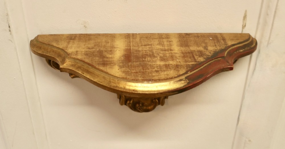 A carved giltwood wall mirror featuring a shell crest hanging directly above a matching gilded wall bracket shelf. View V