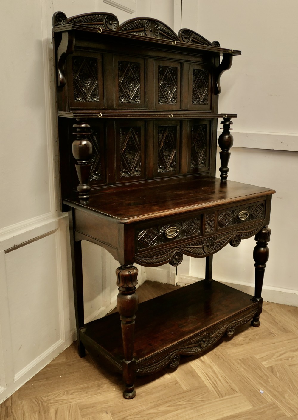 A dark carved oak dresser with tiered upper shelving, geometric diamond-pattern carved panels, two drawers, turned baluster supports, and a wide bottom potboard base. View II