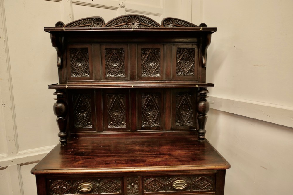A dark carved oak dresser with tiered upper shelving, geometric diamond-pattern carved panels, two drawers, turned baluster supports, and a wide bottom potboard base. View III