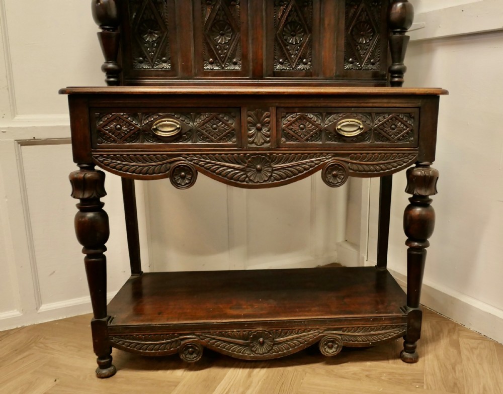 A dark carved oak dresser with tiered upper shelving, geometric diamond-pattern carved panels, two drawers, turned baluster supports, and a wide bottom potboard base. View V