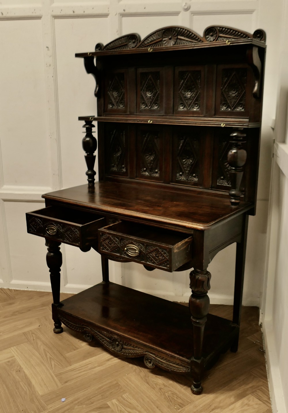 A dark carved oak dresser with tiered upper shelving, geometric diamond-pattern carved panels, two drawers, turned baluster supports, and a wide bottom potboard base. View VI