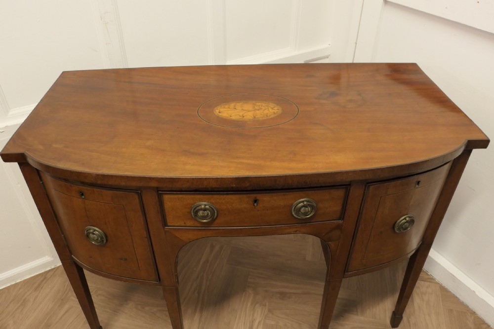 An early nineteenth-century Georgian walnut bowfront sideboard featuring a central drawer, flanking cupboard doors, brass ring handles, central oval wood inlay, and tapered legs. View II