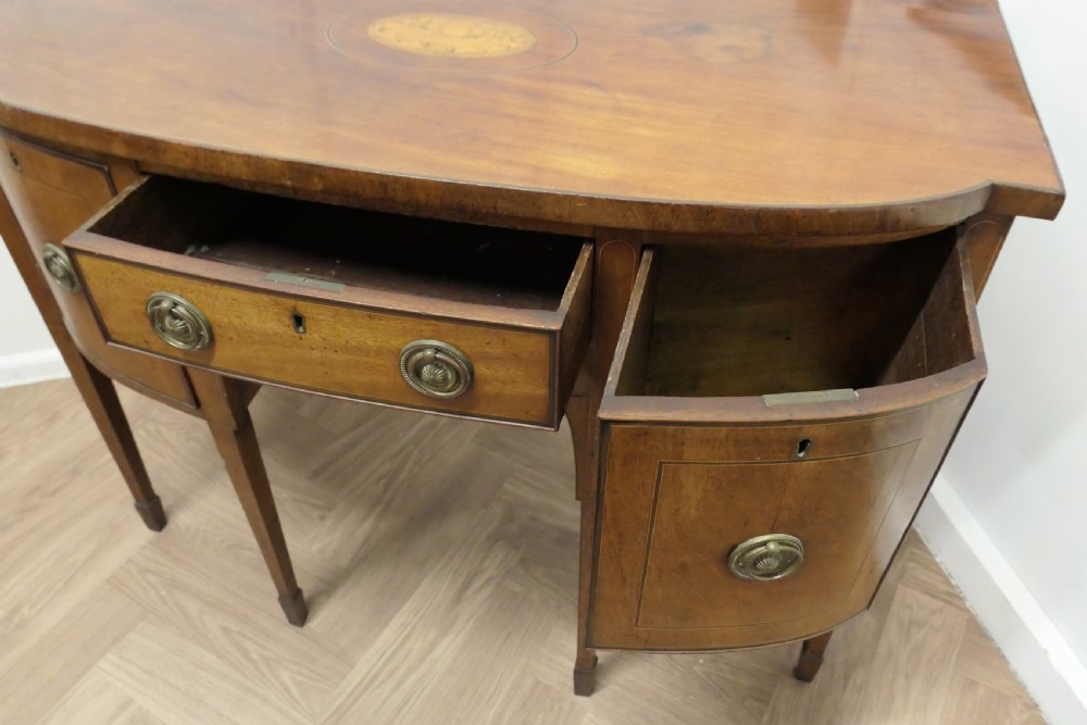 An early nineteenth-century Georgian walnut bowfront sideboard featuring a central drawer, flanking cupboard doors, brass ring handles, central oval wood inlay, and tapered legs. View III