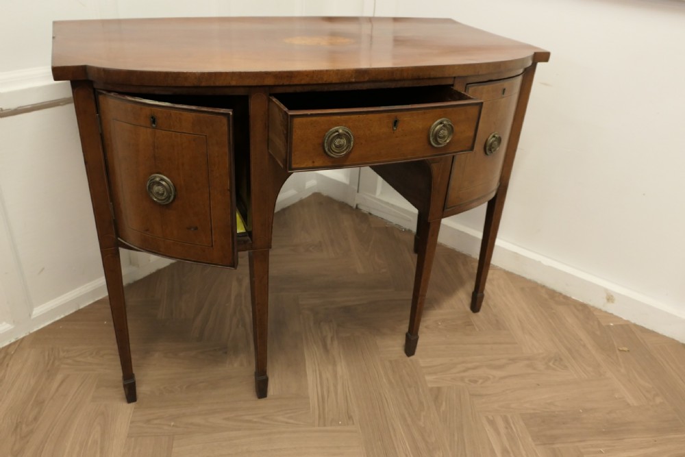 An early nineteenth-century Georgian walnut bowfront sideboard featuring a central drawer, flanking cupboard doors, brass ring handles, central oval wood inlay, and tapered legs. View VI
