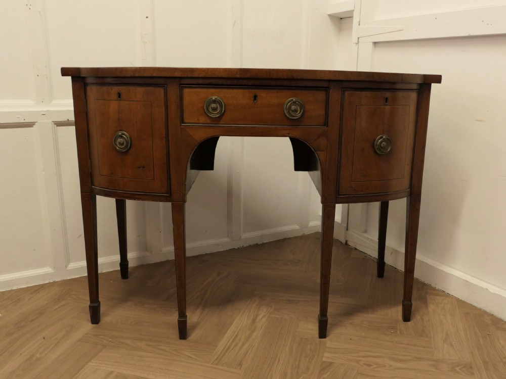 An early nineteenth-century Georgian walnut bowfront sideboard featuring a central drawer, flanking cupboard doors, brass ring handles, central oval wood inlay, and tapered legs. View VII