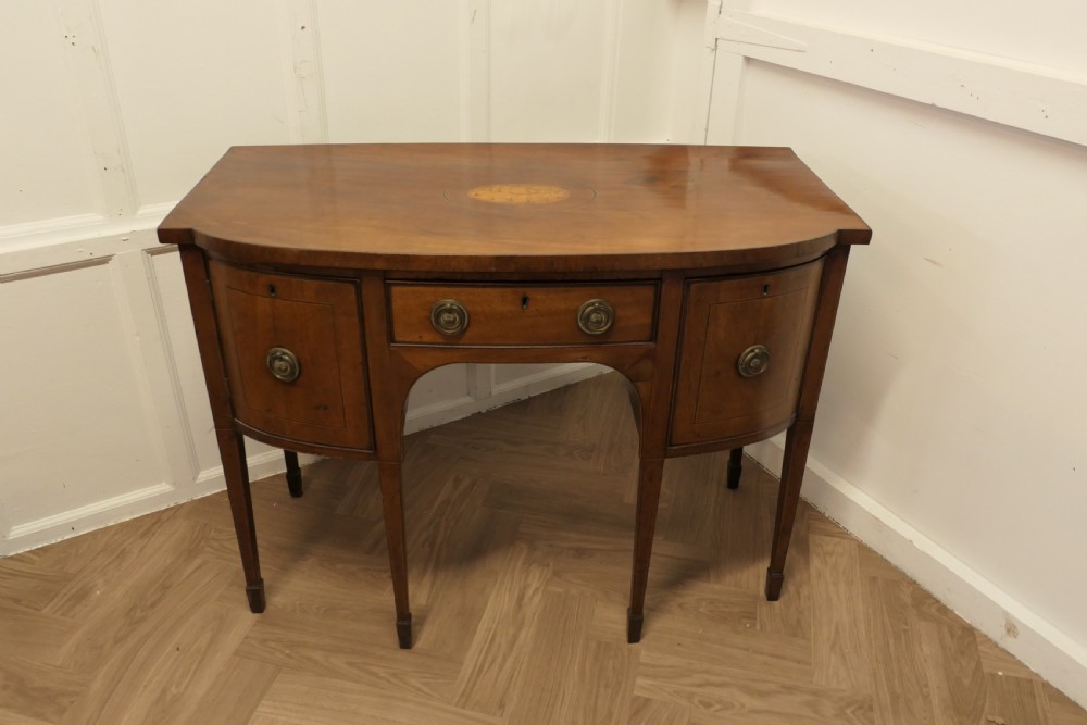An early nineteenth-century Georgian walnut bowfront sideboard featuring a central drawer, flanking cupboard doors, brass ring handles, central oval wood inlay, and tapered legs. View VIII